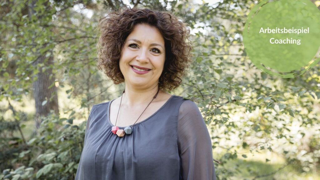 Das Bild zeigt die Autorin, Pia Hübinger. Sie trägt eine graue Bluse und eine Holzkette. Im Hintergrund sind Bäume. Sie lächelt in die Kamera.