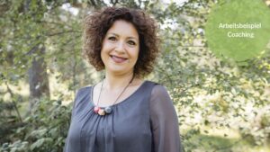 Das Bild zeigt die Autorin, Pia Hübinger. Sie trägt eine graue Bluse und eine Holzkette. Im Hintergrund sind Bäume. Sie lächelt in die Kamera.