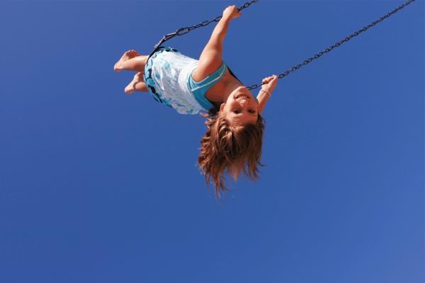 Das Bild zeigt ein Mädchen, das hoch in den Himmel schaukelt. Das Mädchen lacht, seine langen Haare wehen im  Wind. Der Himmel ist strahlendblau. Ein Beispiel dafür, wie wir längst vergessene Freude im Alltag finden können.