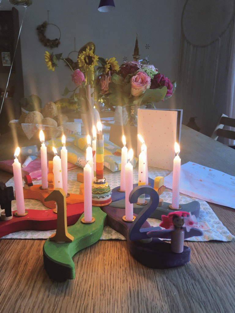 Das Bild zeigt 12 brennende Kerzen in einer Geburtstagssonne. Im Hintergrund stehen Sonnenblumen und Rosen.