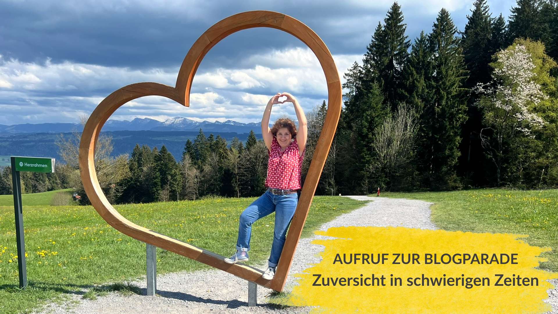 Das Bild zeigt die Verfasserin der Blogparade, Pia Hübinger. Sie steht mit ausgestreckten Armen in einem Metallherz und formt mit ihren Händen ein Herz. Das Bild trägt den Titel: Aufruf zur Blogparade. Zuversicht in schwierigen Zeiten