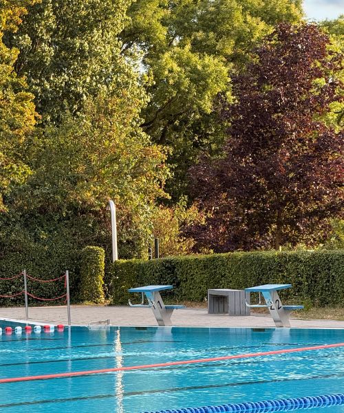 Das Bild zeigt ein Freibad mit türkisfarbenem Wasser, Schwimmerbahnen und Startblöcken. Im Hintergrund stehen Bäume.